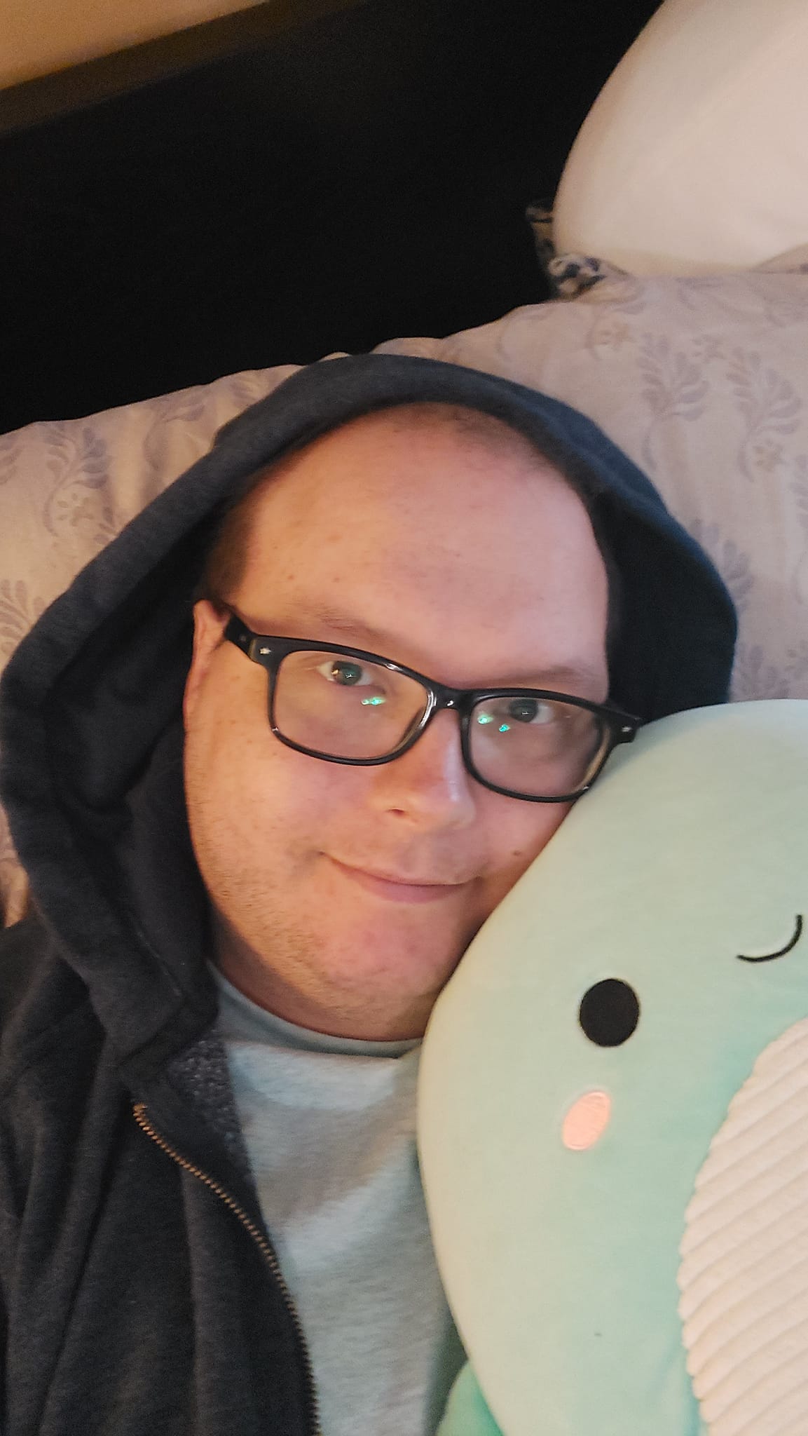 A man lies in bed, fully dressed, holding a Loch Ness Monster Squishmallow, they both look happy